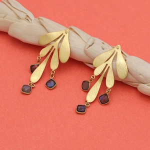 May include: Gold-coloured leaf-shaped earrings with three dangling square-cut gemstones in shades of purple and brown. The earrings are set against a coral background and a white, textured surface.