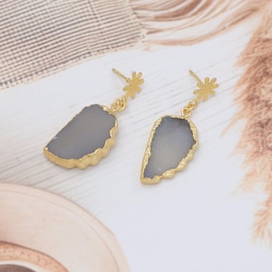 May include: A pair of gold-coloured drop earrings. Each earring features a grey, irregularly shaped stone with a gold-coloured trim. The earrings are suspended from a small, gold-coloured floral stud.