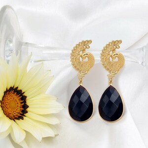 May include: A pair of gold-colored peacock earrings with black teardrop-shaped gems. The earrings feature a detailed peacock design with a textured finish. The gems are faceted, reflecting light. The earrings are displayed on a white background.