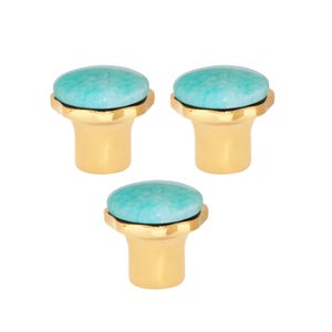 May include: Three cabinet knobs with a gold-coloured base and a round, turquoise-coloured stone top. The knobs have a simple, elegant design, suitable for various furniture styles. The knobs are shown against a white background.