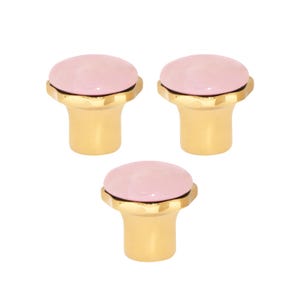 Pode incluir: Três puxadores de armário com base dourada e topo arredondado rosa claro. Os puxadores têm um design simples e elegante, adequados para gavetas ou armários. Os puxadores estão em um fundo branco.