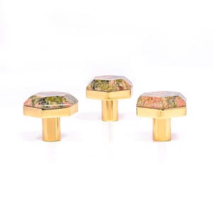 May include: Three cabinet knobs with a gold-coloured base and a faceted, multi-coloured stone top. The stone features shades of green, pink, and brown. The knobs are displayed against a white background.