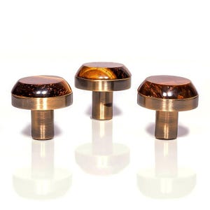 以下が含まれることがあります： キノコ型のデザインの装飾的なキャビネットノブ3つ。各ノブは、茶色と金のタイガーアイ模様のドーム型トップ、真鍮色のバンド、円筒形のステムを備えています。ノブは白い表面に表示されています。