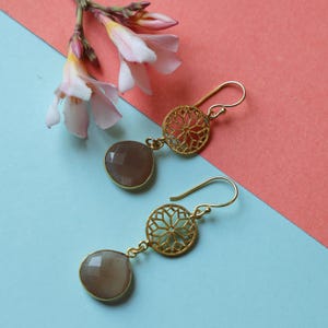 May include: Gold-coloured drop earrings featuring a geometric design and grey, teardrop-shaped gemstones. The earrings are set against a blue and pink backdrop, with a sprig of pink flowers.