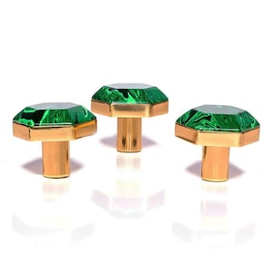 May include: Three decorative cabinet knobs with emerald green faceted tops and gold-coloured bases. The knobs have a geometric design and are arranged against a white background. Suitable for furniture and home decor.