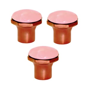 Pode incluir: Três puxadores de armário com um topo rosa arredondado e uma base cor de cobre. Os puxadores têm uma faixa metálica entre o topo rosa e a base. Os puxadores são provavelmente para móveis ou uso decorativo.