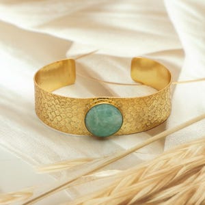 Pode incluir: Uma pulseira de punho dourada com superfície texturizada e uma pedra redonda verde clara. A pulseira tem uma textura martelada e é exibida em uma superfície branca. A pulseira tem cerca de 3 cm de largura.