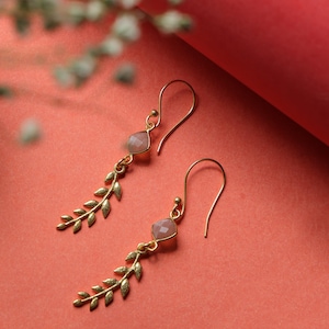 Peut inclure: Paire de boucles d'oreilles pendantes dorées. Chaque boucle d'oreille présente une pierre précieuse rose clair taillée en carré et un motif délicat en forme de feuille. Les boucles d'oreilles sont suspendues à un crochet d'oreille incurvé.
