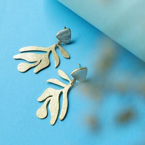 May include: A pair of gold-colored earrings with a leaf-like design. Each earring features a triangular, light-colored stone at the top. The earrings are set against a blue background, creating a striking contrast. These earrings are a stylish accessory.