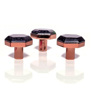 May include: Three geometric cabinet knobs. Each knob has a dark, speckled top and a copper-coloured base and stem. The knobs are set against a white background, offering a clean, modern look. The knobs are suitable for drawers or cabinets.