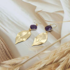 May include: A pair of gold-colored earrings featuring a leaf design. Each earring has a faceted, square-cut, purple gemstone at the top, connected to a textured gold leaf pendant. The earrings are displayed on a neutral background.
