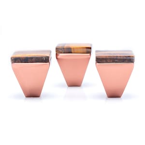 May include: Three copper-coloured cabinet knobs with square tiger's eye stone tops. The knobs have a triangular base. The tiger's eye stone has brown and gold bands. The knobs are set against a white background.