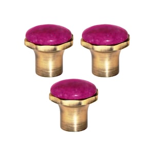May include: Three cabinet knobs with a brass finish and a round, magenta stone top. The knobs have a textured design around the stone. These knobs are suitable for adding a touch of elegance to furniture. The knobs are suitable for furniture.