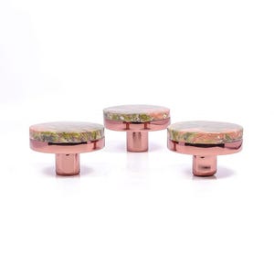 Könnte beinhalten: Drei dekorative Schrankknöpfe mit roségoldener Oberfläche. Jeder Knopf hat eine kreisförmige Oberseite mit einem gesprenkelten grünen, rosa und cremefarbenen Muster. Die Knöpfe sind für Möbel und Heimdekoration gedacht.