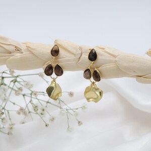 May include: A pair of gold-coloured drop earrings. Each earring has three dark brown, teardrop-shaped stones and a textured, gold-coloured, free-form shape at the bottom. The earrings are displayed on a light-coloured, textured background.