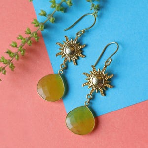 Puede incluir: Pendientes dorados con forma de sol y gemas amarillas y verdes facetadas en forma de lágrima. Los pendientes tienen cierre de gancho y se muestran sobre un fondo azul y rosa.