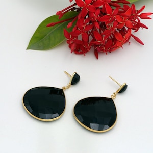 May include: A pair of gold-toned earrings with large, faceted black teardrop-shaped stones. The earrings are displayed with red flowers and green leaves on a white background. The earrings have a post-back closure.