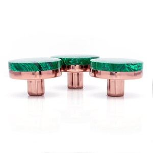 May include: Three cabinet knobs with a green malachite top and a copper-coloured base. The knobs are displayed on a white background. Each knob has a round top and a cylindrical base.