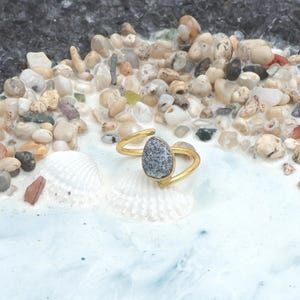 May include: A gold ring featuring a teardrop-shaped grey and white stone. The ring's band curves around the finger, creating an open design. The ring is displayed on a surface with seashells and small pebbles, evoking a beach theme.