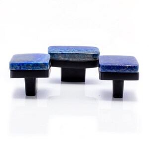 Puede incluir: Tres tiradores cuadrados para armarios con una parte superior de piedra de lapislázuli azul oscuro y una base negra. Los tiradores están dispuestos en fila, cada uno con un patrón diferente de azul y motas doradas.