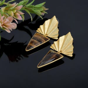 May include: A pair of gold-colored earrings with a fan-shaped top and a triangular, tiger's eye stone. The earrings have a gold-colored frame around the stone and are set against a black background. The earrings are a jewelry accessory.