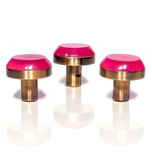 Può includere: Tre pomelli decorativi per mobili a forma di fungo. Ogni pomello ha una parte superiore rosa brillante, una fascia color oro e uno stelo color bronzo. I pomelli sono disposti in fila su sfondo bianco. Per mobili.