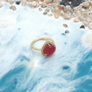May include: A gold ring featuring a teardrop-shaped, red gemstone. The gemstone is encircled by small, clear stones. The ring is set against a blue and white backdrop with seashells and small pebbles.