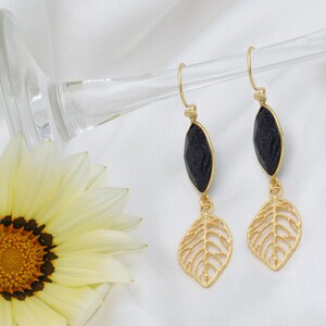 May include: Gold-coloured drop earrings with a dark blue, faceted stone and a gold leaf design. The earrings hang from a simple hook and are displayed against a white background with a yellow flower.
