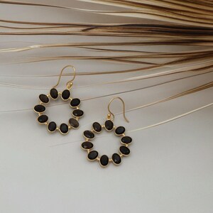 Könnte beinhalten: Ein Paar goldfarbene Creolen, jeweils mit einem Kreis aus dunklen, ovalen Edelsteinen besetzt. Die Ohrringe haben einen einfachen Hakenverschluss und werden vor einem neutralen Hintergrund mit getrockneten Pflanzenelementen präsentiert.