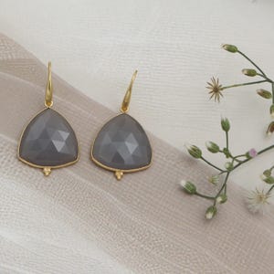 May include: A pair of grey gemstone earrings with a gold-coloured trim and hook. The earrings have a triangular shape with a faceted surface. The background is a neutral colour with a sprig of white flowers.