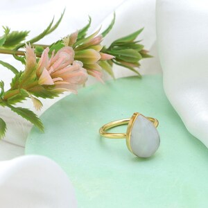 May include: A gold-colored ring with a pear-shaped, light-colored gemstone. The ring is set on a light green surface, with pink and green floral accents in the background. The ring's band is thin and simple.