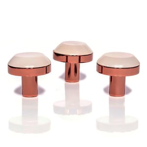 Pode incluir: Três puxadores de armário em forma de cogumelo. Cada puxador tem um topo branco com uma faixa cor de cobre e uma haste cor de cobre. Os puxadores estão dispostos em uma fileira contra um fundo branco.