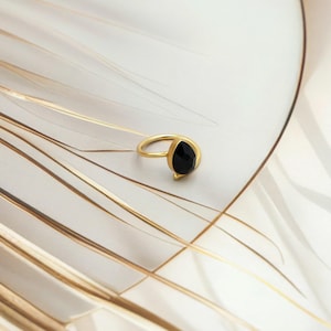 May include: A gold-colored ring with a teardrop-shaped black gemstone. The ring is set on a white and gold patterned surface, creating a contrast between the shiny metal and the dark stone.