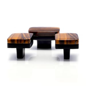 Puede incluir: Tres tiradores decorativos para armarios. Cada tirador presenta una parte superior cuadrada de ojo de tigre con una base y un vástago negros. Los tiradores están dispuestos sobre un fondo blanco, mostrando su diseño y variaciones de color.