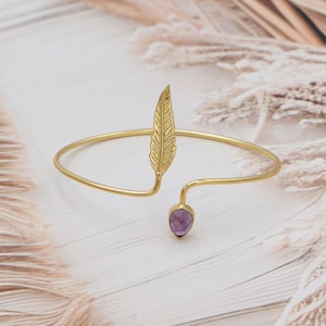 Może przedstawiać: Złota bransoleta na ramię z motywem pióra i fioletowym kamieniem szlachetnym w kształcie łzy. Otwarta konstrukcja ułatwia noszenie. Ten elegancki element jest odpowiedni na różne okazje.