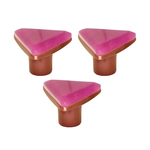 Può includere: Tre pomelli decorativi per mobili. Ogni pomello presenta una parte superiore triangolare rosa traslucida con un bordo color rame e una base cilindrica color rame. I pomelli sono su uno sfondo bianco.