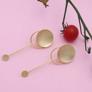 May include: A pair of gold earrings. Each earring has a textured, circular disc connected to a thin, straight bar with a smaller disc at the end. A curved, gold ring joins the disc to the bar. The earrings are set against a pink background.