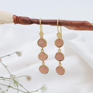 May include: Gold-coloured earrings featuring three peach-hued, irregularly shaped stones. The stones are set in gold bezels and hang from a decorative gold hook. The earrings are displayed on a brown stick against a white backdrop.