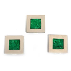 Könnte beinhalten: Drei dekorative quadratische Schrankknöpfe. Jeder Knopf hat einen silberfarbenen quadratischen Rahmen mit einem leuchtend grünen, gewirbelten, quadratischen Einsatz. Die Knöpfe sind vor einem weißen Hintergrund angeordnet.