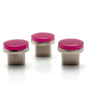 May include: Three cabinet knobs with a bright pink, circular top and a brushed silver base. The knobs have a modern, minimalist design, perfect for updating furniture. The knobs are shown against a white background.