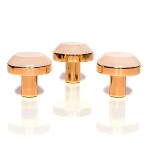 Pode incluir: Três objetos decorativos em forma de cogumelo. Cada um tem uma haste dourada e um topo arredondado com uma superfície rosa claro e uma borda dourada. Os objetos estão dispostos em uma fileira contra um fundo branco.