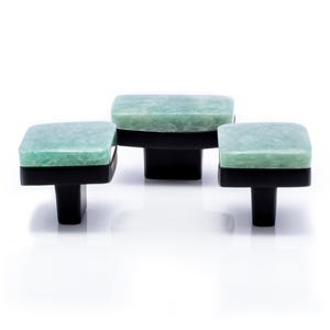 May include: Three square cabinet knobs with a light green, speckled surface and black bases. The knobs are arranged in a staggered pattern. Each knob has a T-shaped base. The knobs are suitable for cabinets.