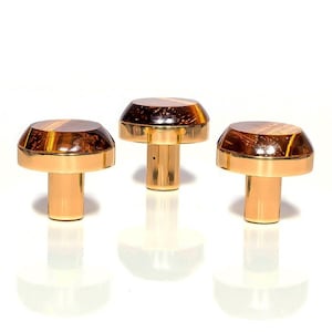 May include: Three decorative bottle stoppers. Each stopper has a gold-colored base and a domed top with a brown and amber pattern. The stoppers are arranged on a white surface, with their reflections visible.