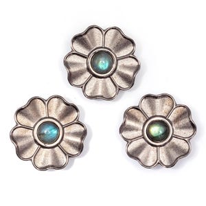 Könnte beinhalten: Drei silberfarbene, blütenförmige Dekorationsgegenstände mit einem zentralen, irisierenden, runden Stein. Die Blütenblätter haben eine geriffelte Textur. Die Steine haben eine blau-grüne Farbe. Die Objekte sind auf weißem Hintergrund angeordnet.