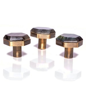 May include: Three cabinet knobs with a geometric design. Each knob has a dark, faceted top, a gold-coloured band, and a cylindrical base. The knobs are arranged on a white surface, with their reflections visible.