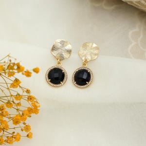 Pode incluir: Um par de brincos dourados com uma pedra central preta facetada. Os brincos têm uma peça superior texturizada de formato irregular e um halo de pequenas pedras transparentes ao redor da pedra preta. Apresentados em tecido creme.