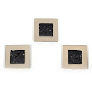 Könnte beinhalten: Drei quadratische, silberfarbene Untersetzer mit einem dunklen, gesprenkelten Zentrum. Die Untersetzer sind vor einem weißen Hintergrund angeordnet. Jeder Untersetzer hat eine kleine, gebogene Lippe am unteren Rand.