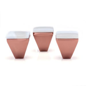 May include: Three geometric containers with a rose gold finish and white tops are displayed against a white background. The containers have a diamond-like shape, with a square top and a pointed bottom. The white tops provide a contrast to the metallic finish.