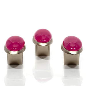 May include: Three cabinet knobs with a contemporary design. Each knob has a bright pink, rounded glass or acrylic top set on a brushed metal base. The knobs are displayed against a white background.