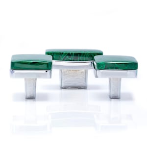 May include: Three cabinet knobs with square, malachite green tops and silver-toned bases. The knobs are arranged in a row, with the centre knob slightly larger. The green tops have a marbled appearance, and the bases are rectangular.